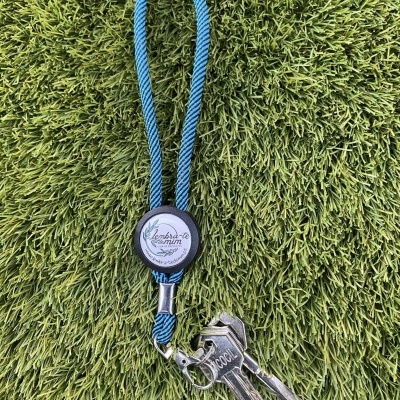 Chaveiro com cordão azul, etiqueta preta com texto e três chaves sobre relva verde