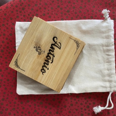 Caixa de madeira com texto Antonio e saco de tecido branco sobre pano vermelho com padrão floral