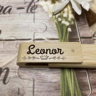 Clipe de madeira com nome 'Leonor' gravado, sobre puzzle com padrão de madeira e flores brancas
