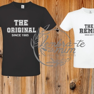 Duas t-shirts penduradas em cabides, uma preta e outra branca, com textos impressos na frente, em fundo de madeira rústica