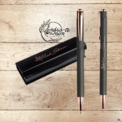 Dupla de canetas cinzentas com detalhes rosa dourado e caixa preta com texto em rosa dourado sobre madeira clara