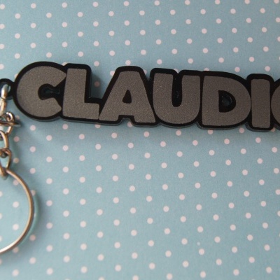 Porta-chaves com o nome CLAUDIO em letras cinzentas com contorno preto sobre fundo azul claro com bolinhas brancas.