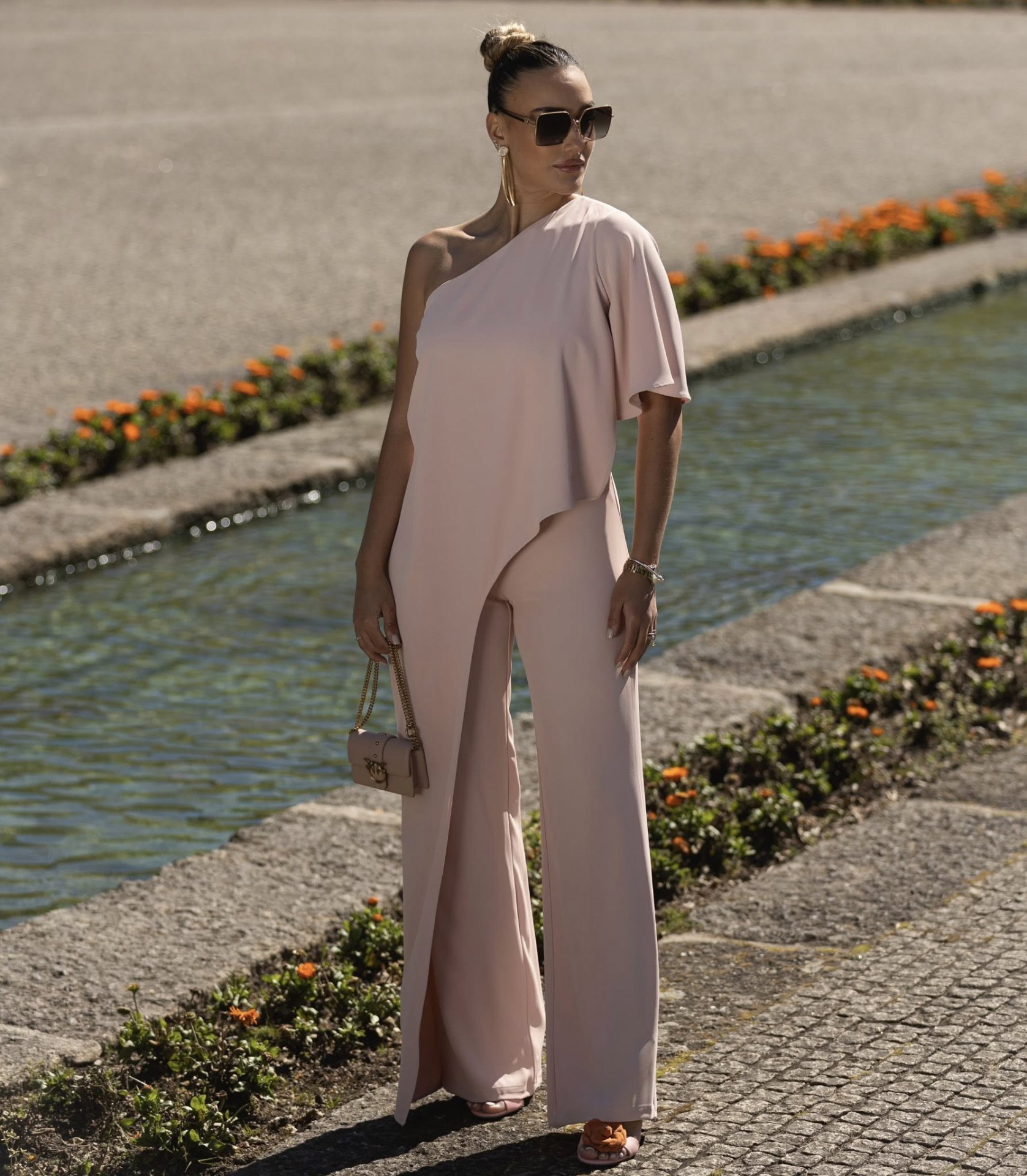 Macacão assimétrico rosa com folho Miss You Mulher com macacão rosa claro de uma manga só e óculos escuros perto de canal de água