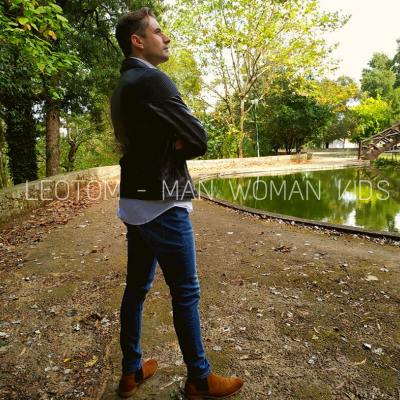 Homem de perfil com blazer preto e calças de ganga azul junto a lago ao ar livre