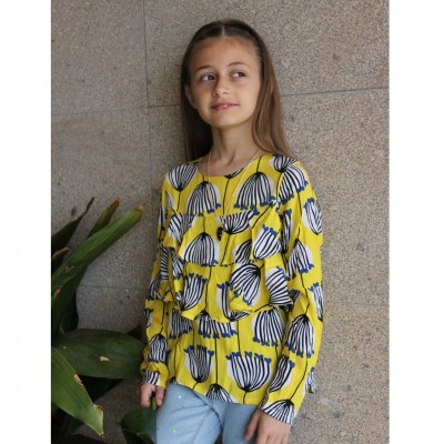 Menina com blusa amarela estampada e calças azuis junto a parede cinza e plantas