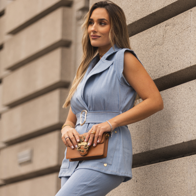 Mulher com colete e calças de ganga azul claro, segurando mala castanha com ornamentos dourados e texto Just Cavalli Milano, junto a parede de pedra cinza.