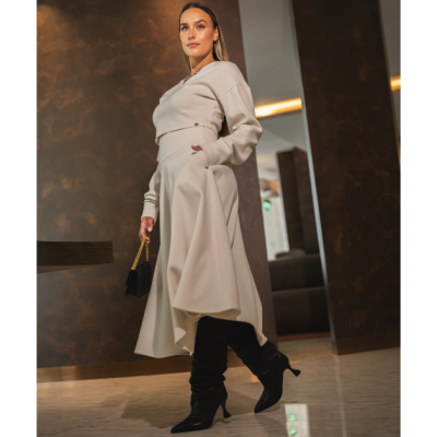 Conjunto feminino bege com top de manga comprida e saia assimétrica fluida, sapatos pretos de bico fino com salto médio e mala preta pequena com alça dourada.