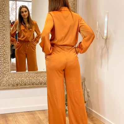 Conjunto de roupa laranja feminino com camisa e calças em espaço interno com espelho e chão de madeira.