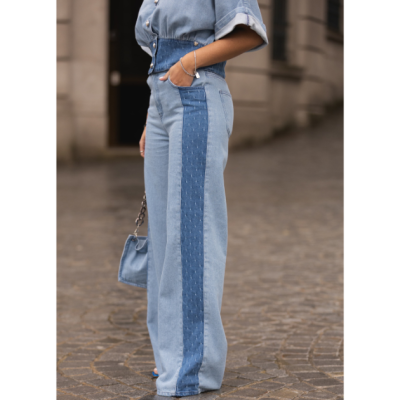 Calças de ganga largas azuis com faixa azul escura e costuras, camisa de ganga azul clara, mala azul pequena com alça de corrente num pavimento de pedra