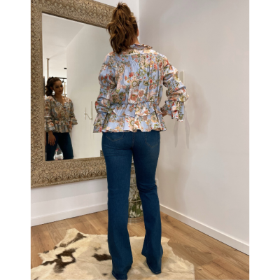 Mulher de costas usando blusa floral com babados e calças jeans azuis em pé diante de espelho grande em sala com chão de madeira e tapete claro com manchas escuras