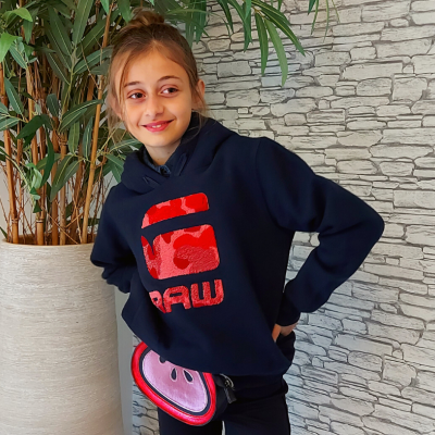 Menina com hoodie azul e bolso vermelho em ambiente com planta e parede decorativa cinzenta