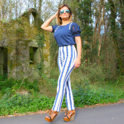 Mulher com blusa azul e calças de riscas azuis e brancas numa estrada ao lado de vegetação e ruína