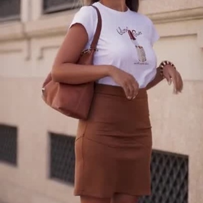 Mulher com t-shirt branca, saia castanha e mala a caminhar numa rua com texto miss you