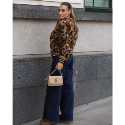 Mulher com casaco animal print, calças largas de ganga e bolsa bege junto a parede cinzenta