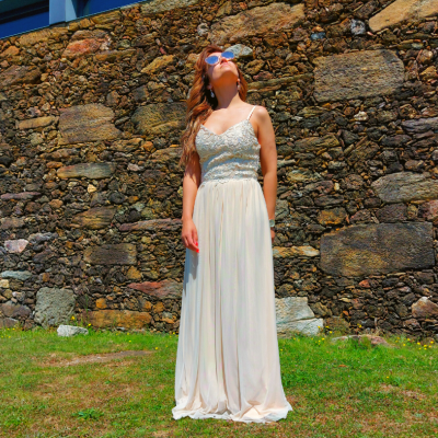 Mulher de vestido longo branco com corpete de renda azul clara em frente a muro de pedra