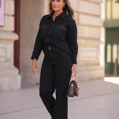 Mulher com camisa preta com tachas, calças pretas largas, sapatos castanhos e mala Louis Vuitton castanha com padrão monograma.
