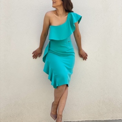 Vestido azul turquesa de um ombro com babados, mulher de sapatos nude encostada numa parede branca