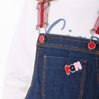 Macacão de ganga azul escuro com suspensórios de riscas vermelho e rosa sobre t-shirt branca.