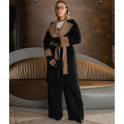 Mulher com casaco preto e castanho em ambiente interior com sofá e parede castanha.