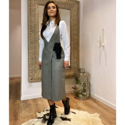 Mulher com camisa branca e vestido cinza risca de giz em ambiente interno com piso de madeira e espelho grande.