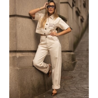 Mulher a usar conjunto creme com camisa curta e calças compridas com tachas, sapatos castanhos metálicos e óculos escuros pretos junto a parede de pedra.