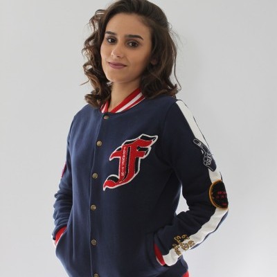 Mulher de casaco bomber azul escuro com riscas vermelhas e brancas e patches coloridos, posed contra fundo branco.