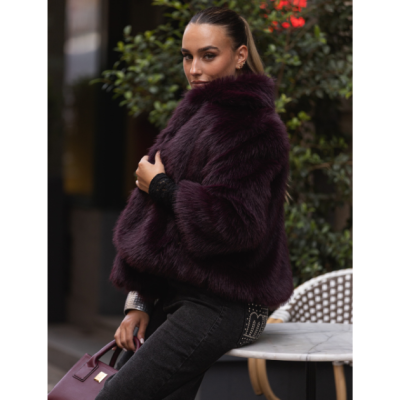 Mulher veste casaco curto de pelo bordô, calças pretas e segura mala bordô ao lado de mesa exterior