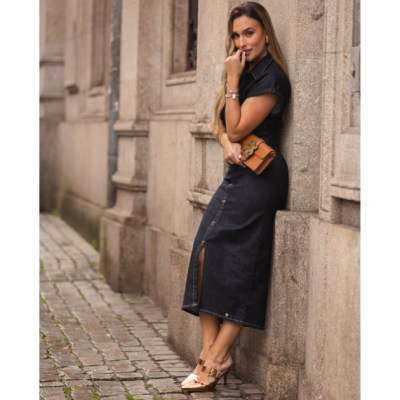 Mulher com vestido preto comprido em ganga, mala castanha e sapatos castanhos junto a parede de pedra num passeio de calçada de cubos.