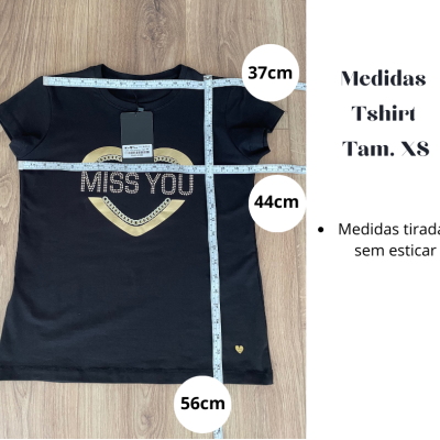 T-shirt preta com estampado de coração amarelo e texto branco 'MISS YOU', medidas indicadas por fita métrica e etiquetas penduradas.