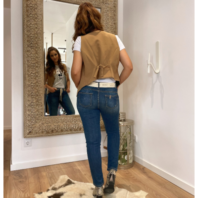 Mulher viste colete castanho, t-shirt branca, jeans azuis e botas com padrão de cobra diante de um espelho grande com moldura trabalhada