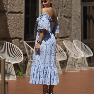 Mulher com vestido azul claro brocado com folhos e sandálias de cunha, em fundo com parede de pedra e cadeiras brancas de metal