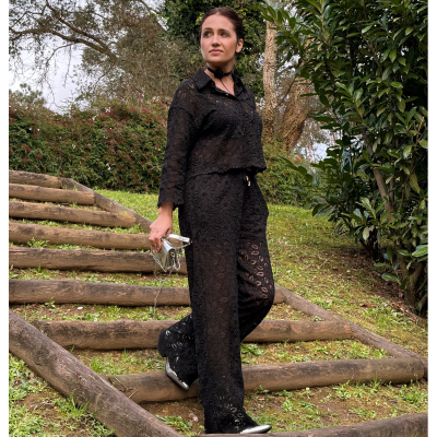 Conjunto preto rendado feminino com calça e camisa, ténis pretos e bolsa prateada, cenário ao ar livre com escadas de madeira e vegetação.