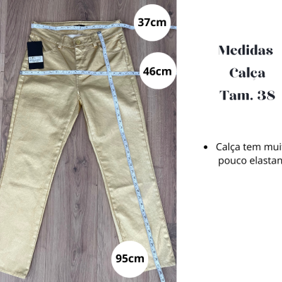 Calça dourada metálica sobre chão de madeira com fita métrica e texto de medidas junto