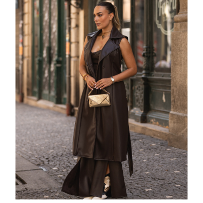 Mulher com colete longo castanho escuro e mala dourada numa rua antiga com calçada de pedra