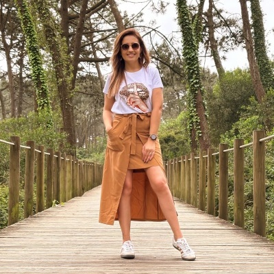 Mulher com saia midi castanha, t-shirt branca estampada e ténis brancos na passadeira de madeira
