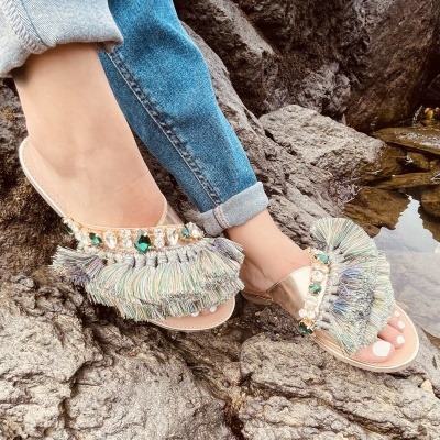 Sandálias decorativas com franjas e pedras em ambiente rochoso junto a água