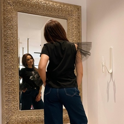 Mulher veste t-shirt preta com detalhe de tule, jeans azuis junto a espelho com moldura dourada