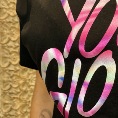 T-shirt preta com estampado em letras rosa, branco, azul e verde em fundo bege texturizado