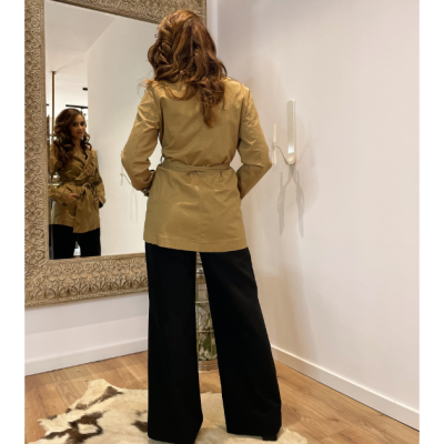 Mulher de costas usa casaco bege com cinto e calças pretas largas em sala com espelho e tapete.