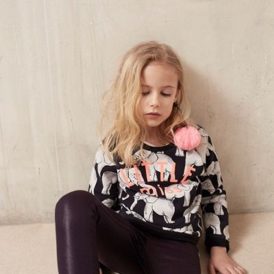 Menina com casaco estampado de cavalos e calças púrpuras sentada no chão junto a parede cinza.