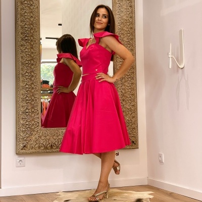 Mulher com vestido rosa forte e sandálias douradas em frente a um espelho grande com moldura trabalhada