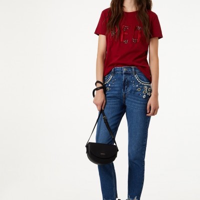 Mulher com t-shirt vermelha, jeans azuis com aplicação de pedras e botas pretas com tachas, segurando mala preta pequena