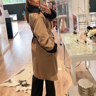 Mulher com casaco castanho e detalhes pretos a tirar selfie numa loja moderna com chão de madeira.