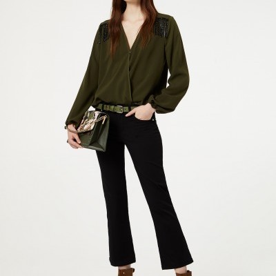 Mulher com blusa verde, calças pretas, botins castanhos e mala verde em fundo branco