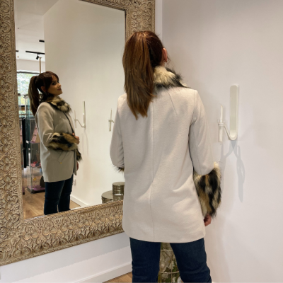 Mulher de costas a usar casaco bege com detalhes em pelo, em frente a um espelho grande com moldura decorativa