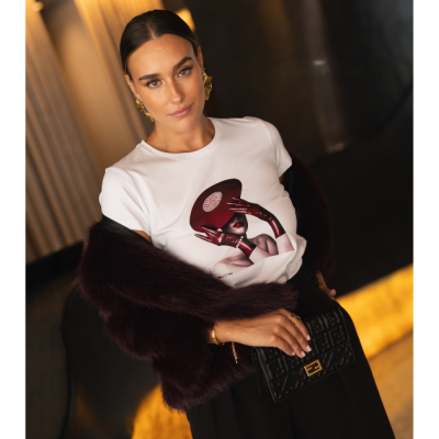 Mulher com t-shirt branca estampada, casaco vinho, mala preta e fundo interior iluminado