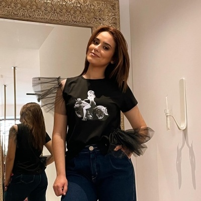 Mulher com t-shirt preta com mangas de tule e estampa prateada, jeans azuis, junto a um espelho com moldura dourada