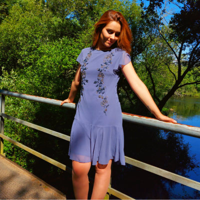 Mulher a usar vestido lilás com bordados florais junto a um rio num dia soalheiro