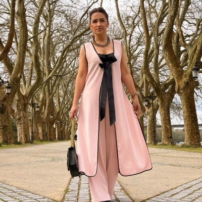 Conjunto feminino rosa claro com casaco longo, laço preto e mala preta em ambiente exterior