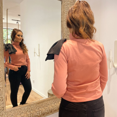 Mulher em casaco rosa com detalhe preto no ombro em frente a espelho com moldura branca